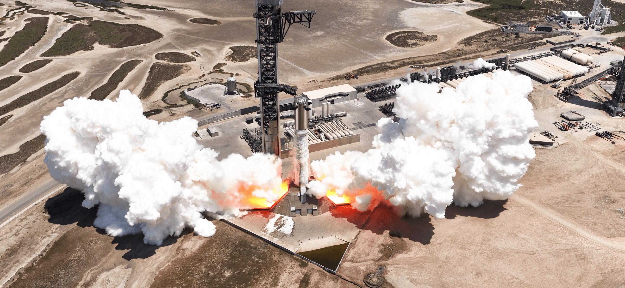 Overhead view of Starship on Pad 2 at Starbase during a static fire, white steam and flame over the coastal industrial site. Official SpaceX photograph.