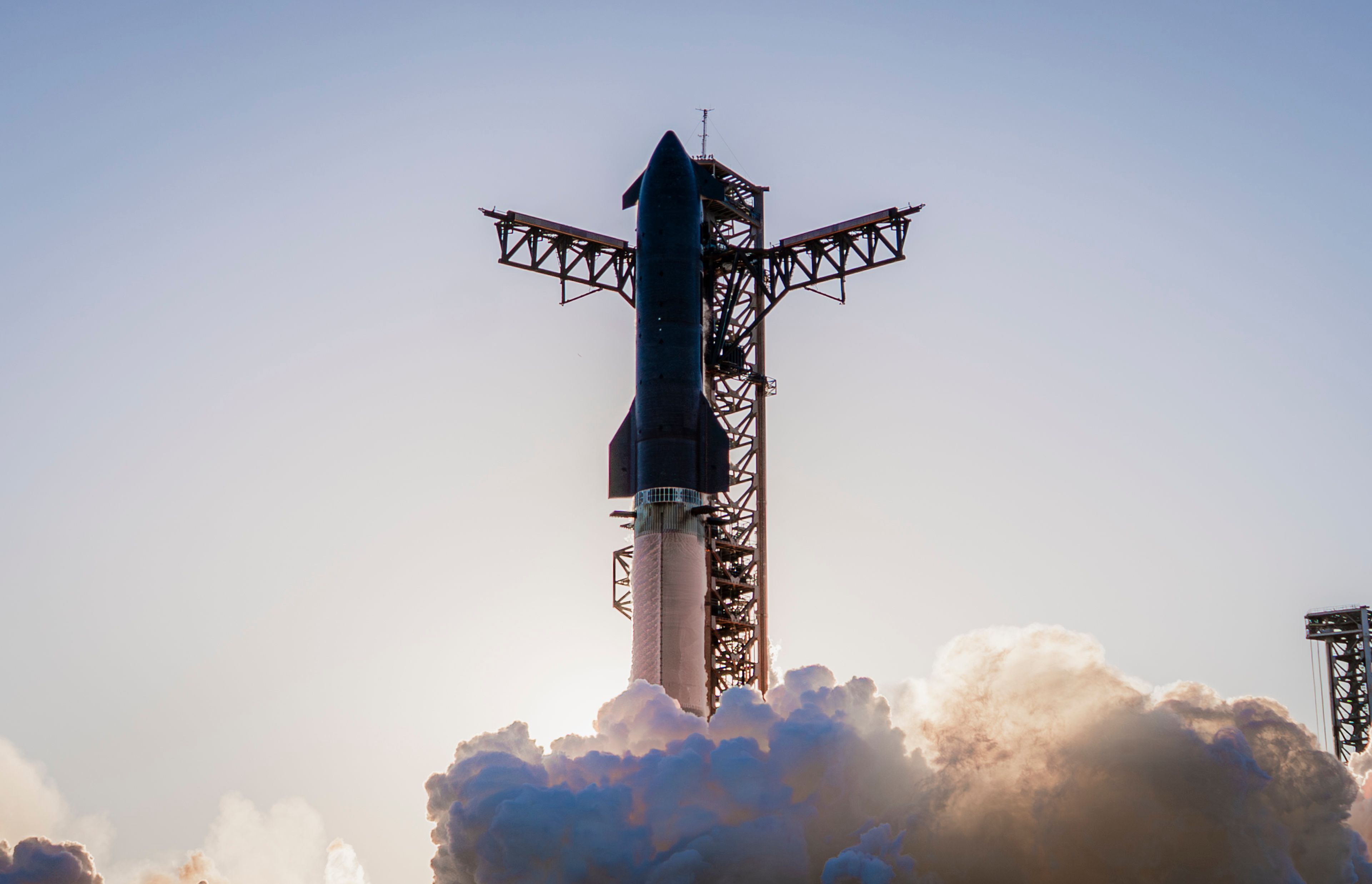 Starship and Super Heavy lifting off from the orbital launch mount at Starbase, chopsticks open, plume filling the pad. Official SpaceX photograph.