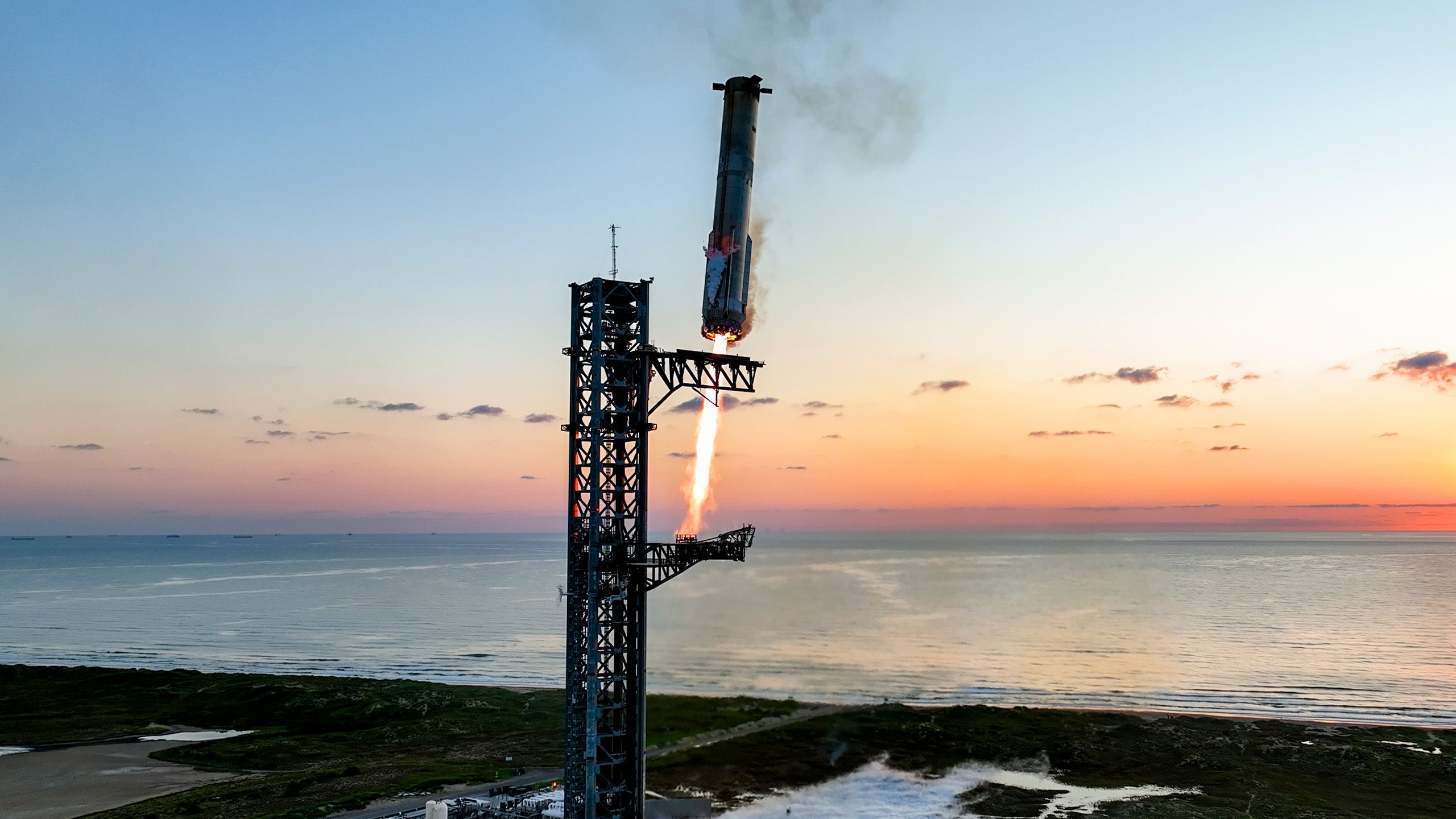 Starship caught at the tower over the Gulf at sunset, chopsticks closing on the vehicle. Official SpaceX photograph.